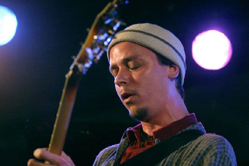 Guitarist Kurt Rosenwinkel performing in a knit cap, photographed in profile under blue-toned stage light.